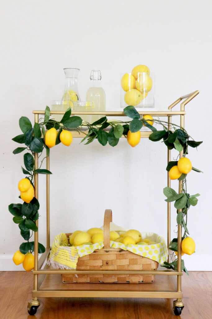 Drink cart in the middle of photo lined with a lemon garland, basket of lemons on the bottom shelf, and lemonade, cookies and a vase of lemons on the top.