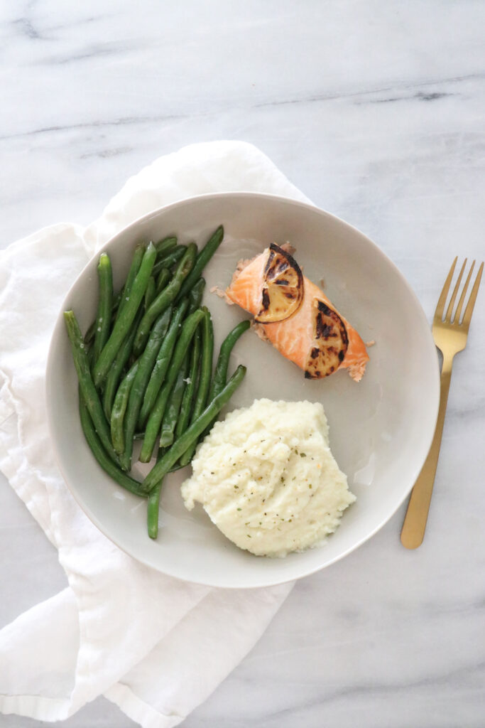 White plate in center of photo with salmon, mashed cauliflower and green beans all on white napkin. Gold fork to the right of plate.