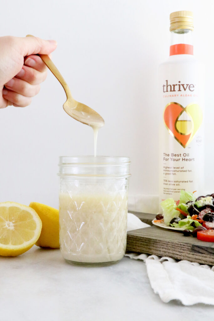 Lemon Tahini Dressing in center of photo with spoon in glass container. Half of lemon next to dressing. Thrive algae oil in picture and salad taco on the side.