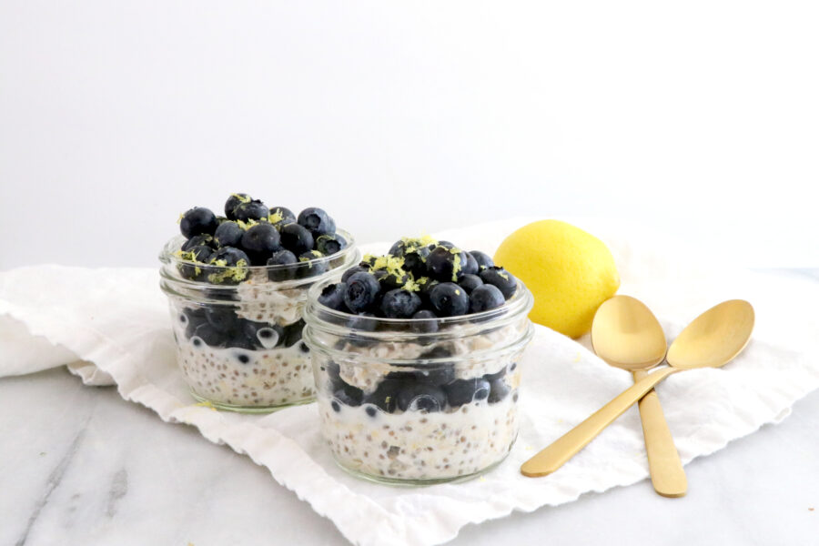 Two glass jars filled with layers of overnight oats and fresh blueberries. Two gold spoons on the side with a lemon.
