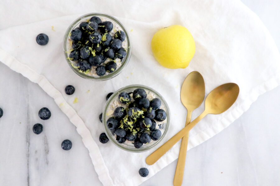 Two glass jars filled with layers of overnight oats and fresh blueberries. Two gold spoons on the side with a lemon. Blueberries on the left side.