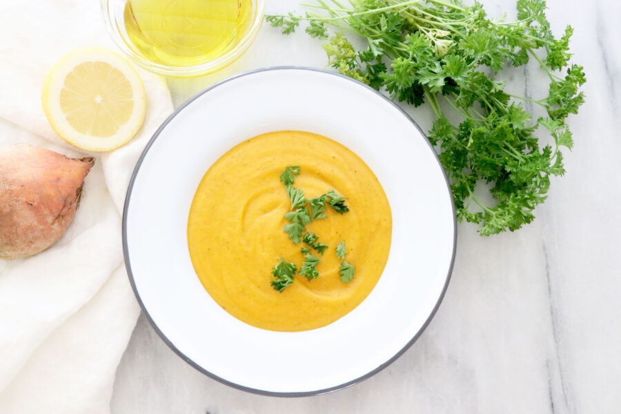 White bowl with creamy vegetable soup topped with parsley surrounded by onion, lemon half, olive oil and parsley.