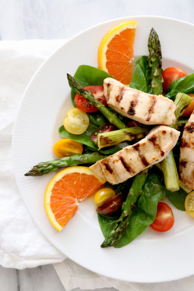 White plate of salad topped with asparagus, orange slices, tomatoes and grilled chicken. 