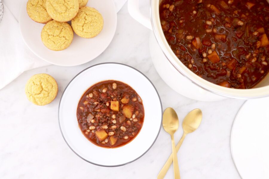 White bowl of lentil chili and plate of corn muffins in left corner. Pot of soup on the right and two gold spoons in bottom right corner.