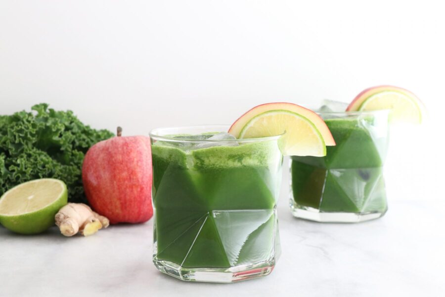 Two glasses filled with green juice and fruit and veggies in the background.