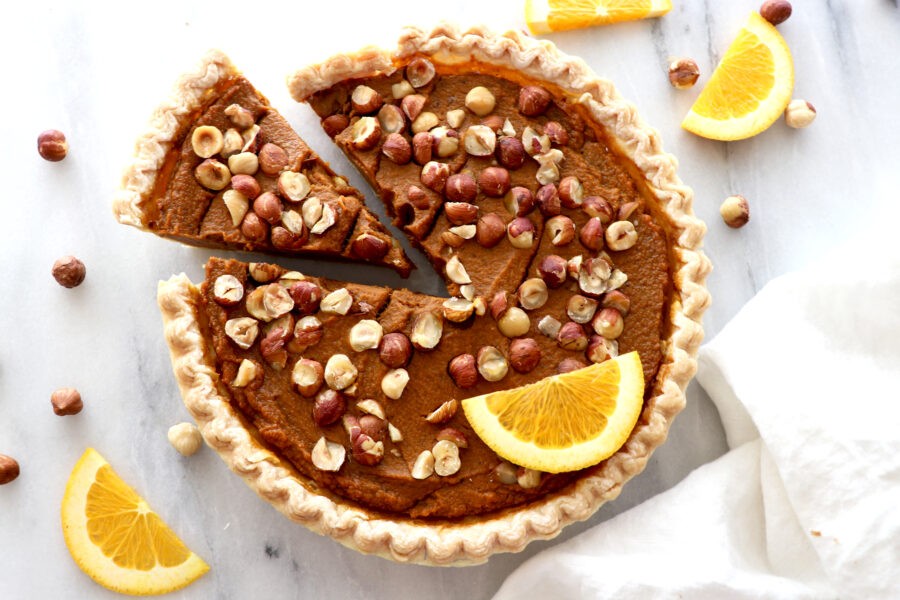 Orange hazelnut pumpkin pie topped with hazelnuts, orange slice and slice cut out. Surrounded by hazelnuts and orange slices.