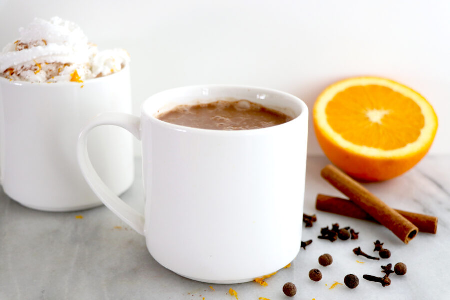 White mug with orange spiced hot coco. Surrounded by orange slice and spices.