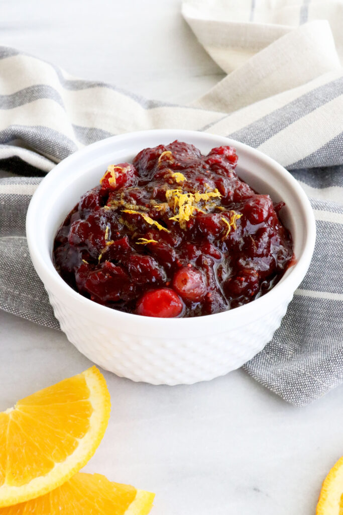 White bowl filled with orange cranberry sauce surrounded by napkin and slices of orange.