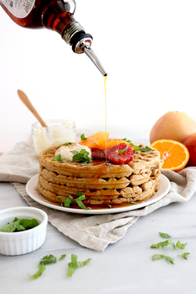 White plate with winter citrus spiced waffles being topped with maple syrup and winter fruit. Citrus around photo.