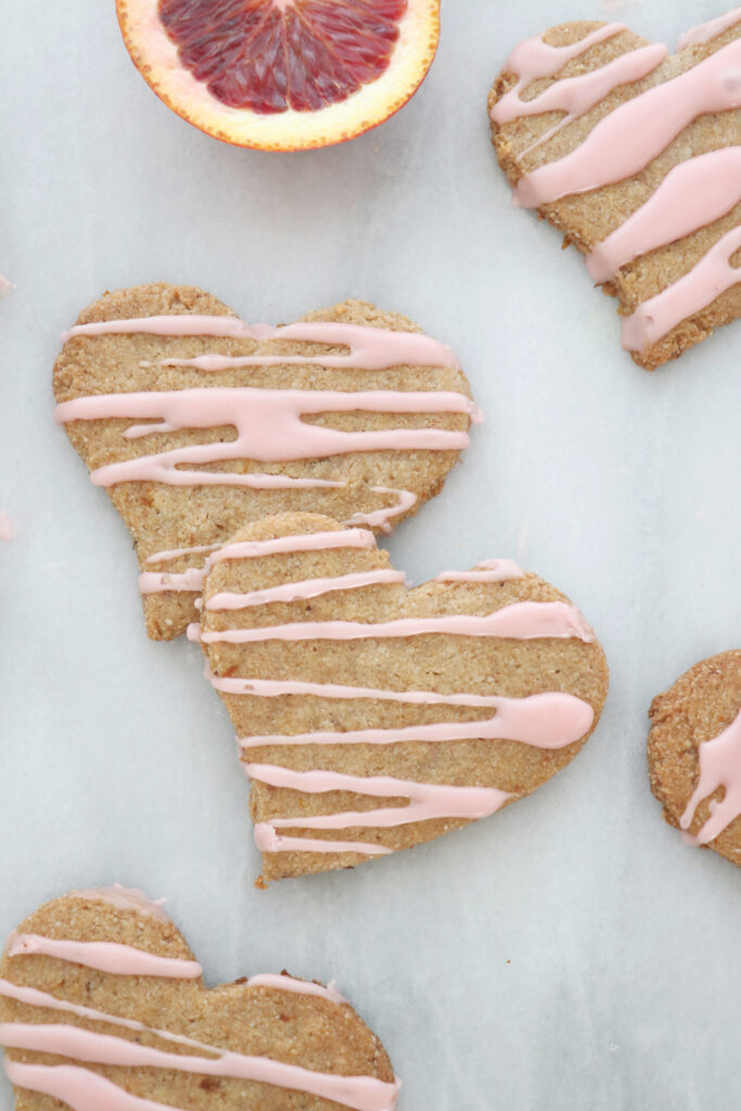Four heart shaped cookies with pink icing on top. Slice of blood orange in background.