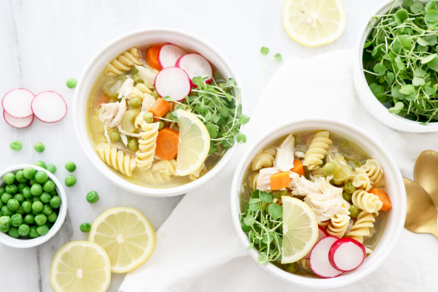 Two bowls of lemon chicken soup filled with veggies and vegetable slices surrounding bowls.