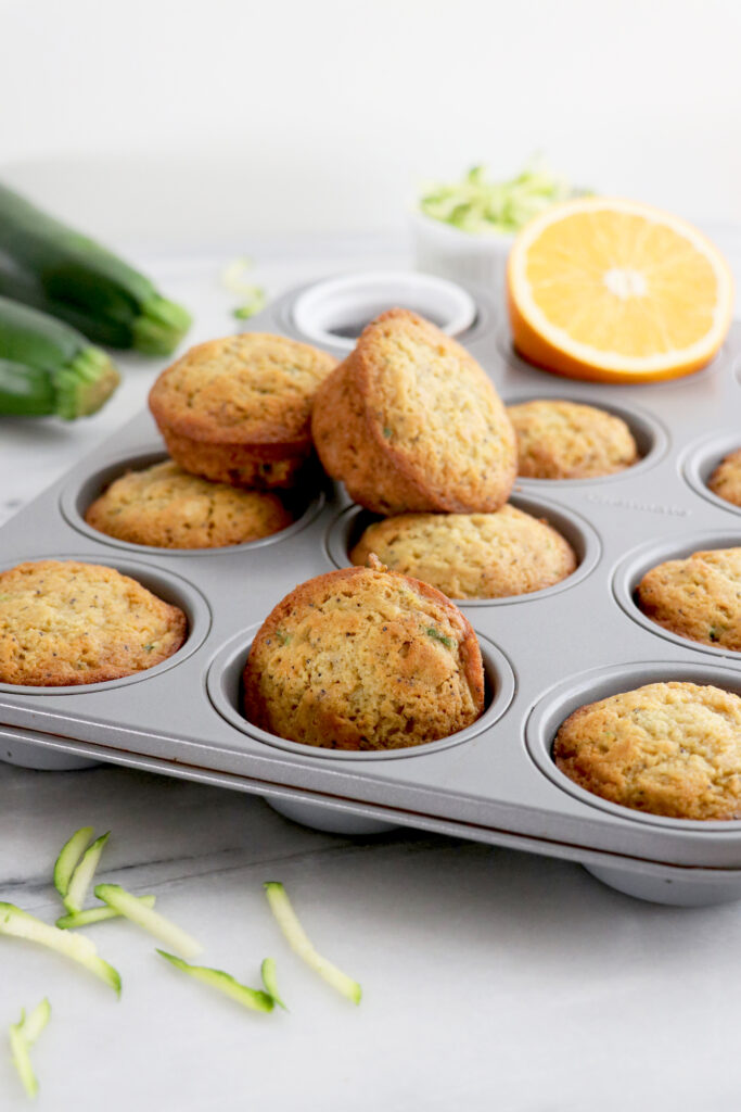 Orange zucchini muffins in silver muffin tin with two on top and orange slice on the side.