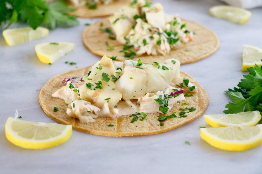 Two corn tortillas topped with citrus fish, homemade slaw. Surrounded by lemon slices and cilantro.