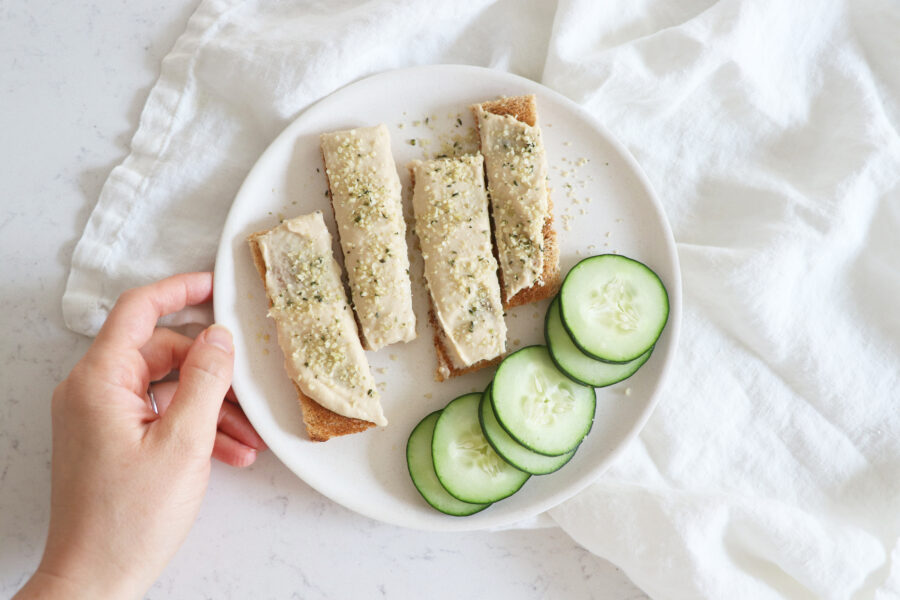 White plate with toast sliced topped with hummus and sliced cucumber.