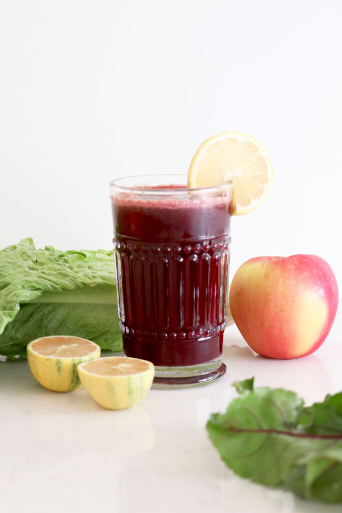 Glass of beet juice with lemon slice on top. Juice surrounded by produce.
