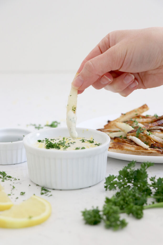 White bowl with lemon aioli and hand dipping in parsnip fries.