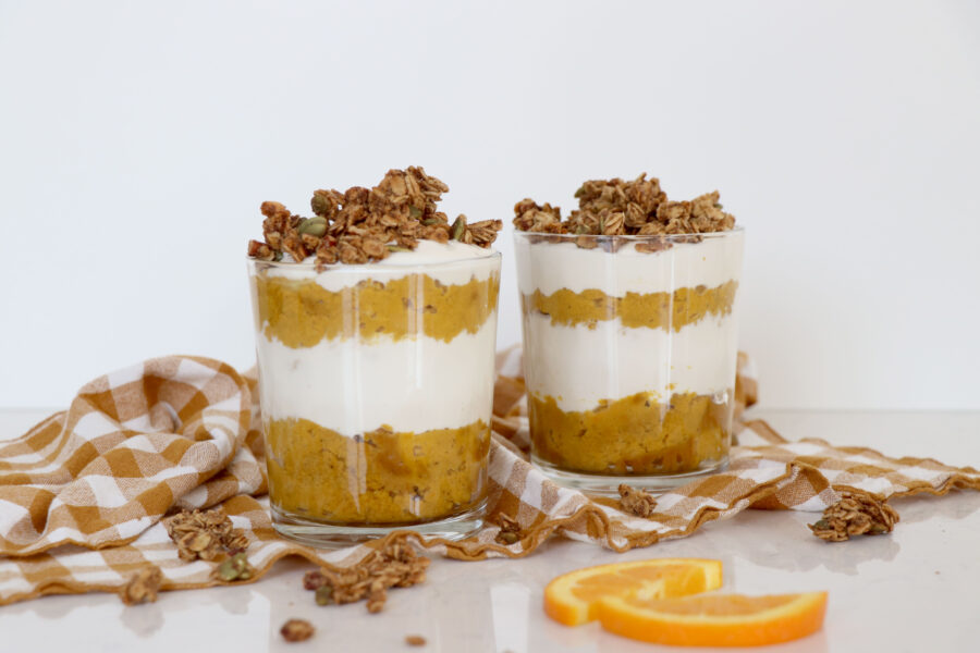 Two glasses layered with pumpkin pie parfait and topped with granola. Brown checkered towel in background.