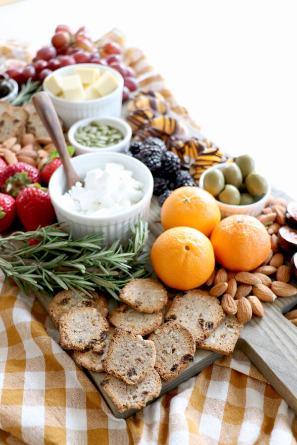 Wooden board with oranges, bread, nuts, crackers, fruit and dips.
