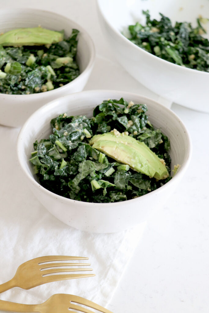 3 white bowls of kale salad with sliced avocado.